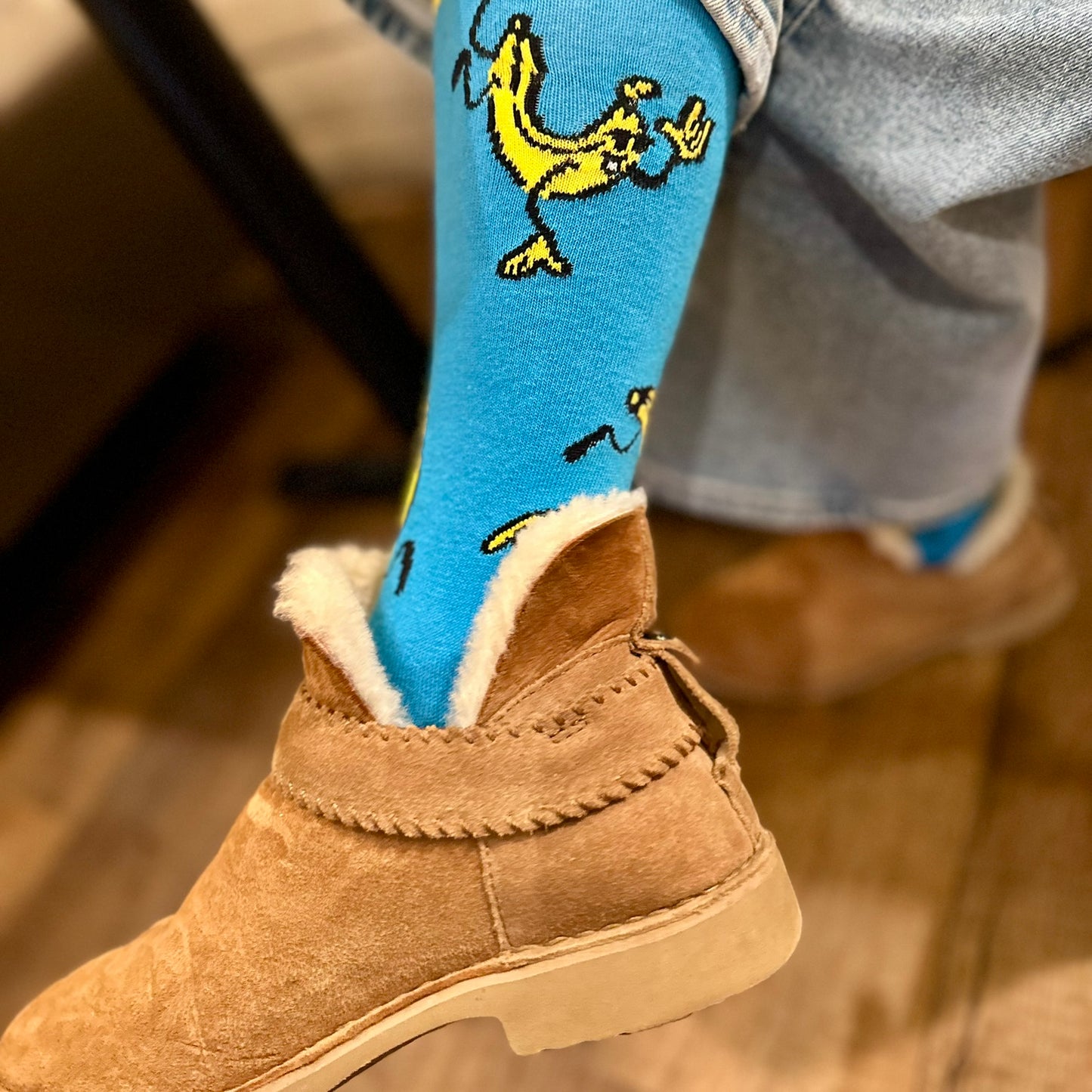 Person wearing blue socks with banana design and brown boots on a wooden floor.