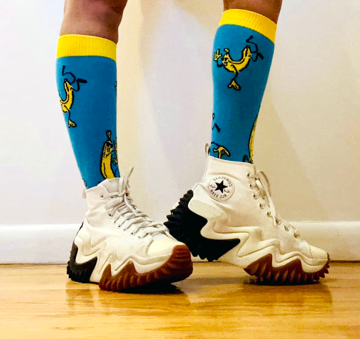 White sneakers with black soles worn with blue socks featuring yellow accents and black designs on a light wooden floor.