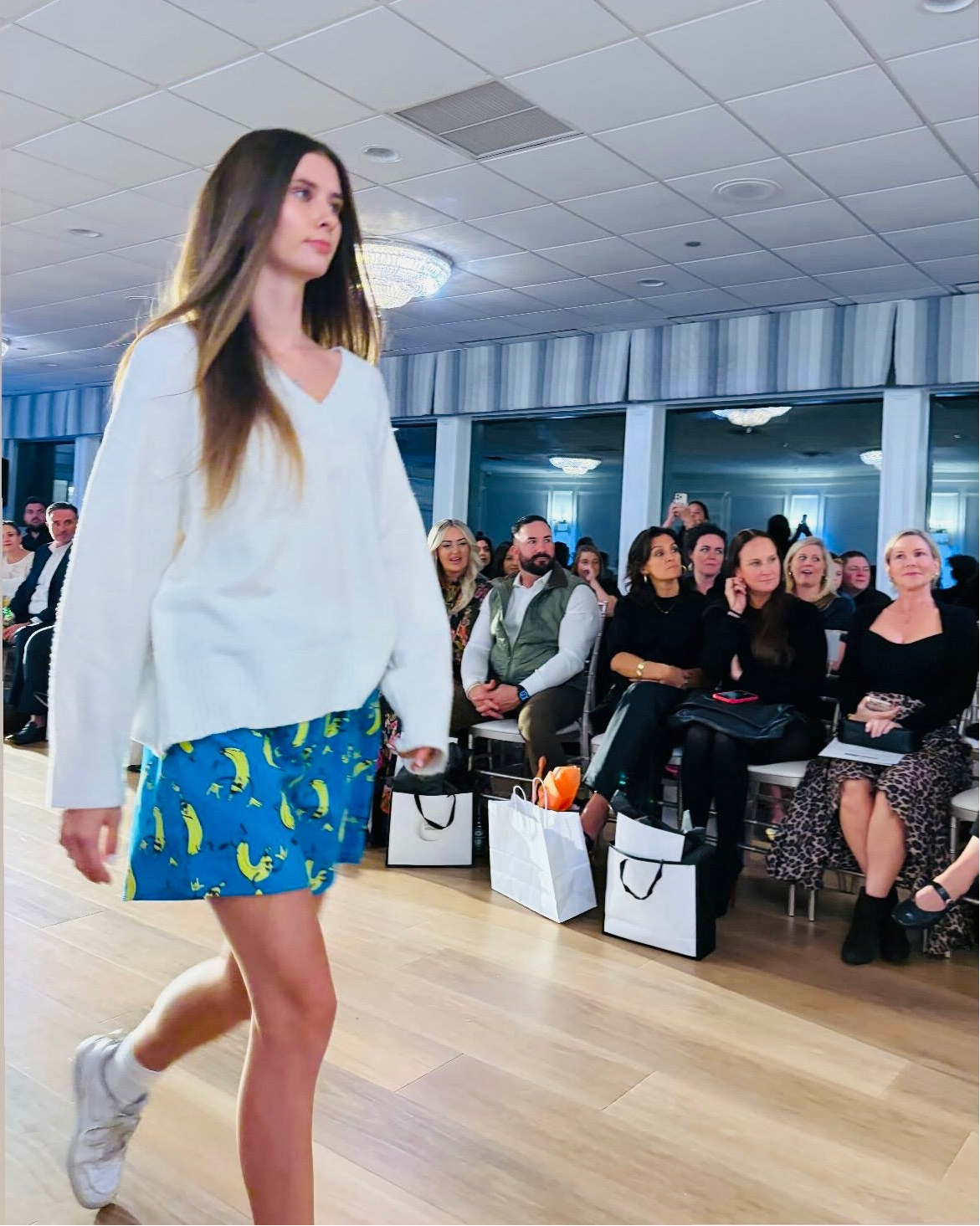 Model walking on a runway wearing a white sweater and blue skirt with banana pattern, audience seated in the background.